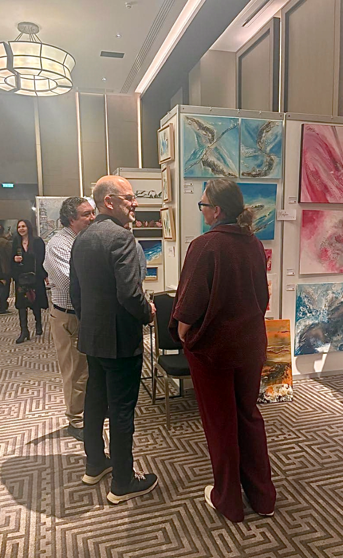 Visitors engaging with abstract mixed-media artworks at ART.CY 2026 exhibition, Amara Hotel Limassol, including coastal textured paintings in blue and pink tones